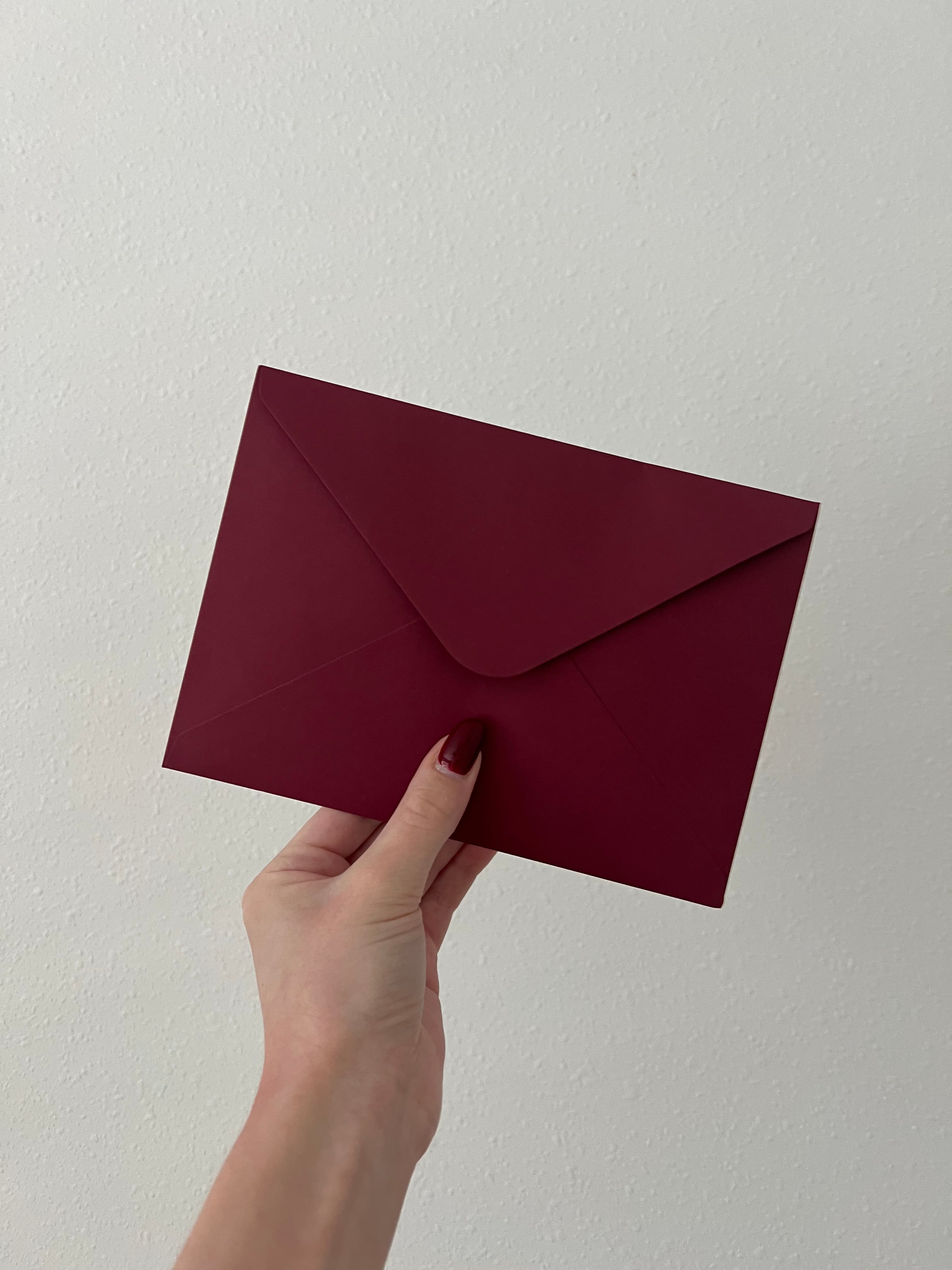 A hand holding a burgundy Letters of Faith envelope against a cream wall